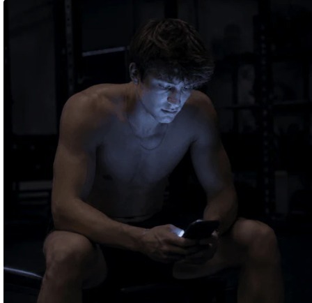 Lean athlete sitting alone on a gym bench in near-darkness, face and chest lit only by the cold blue glow of his phone